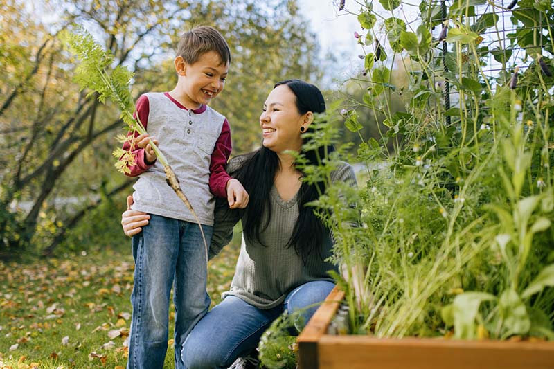2025-07-16-acrf-home-find-alaska-native-resources-800x533px-4q An adult woman kneels down next to a child who is holding a carrot in a garden